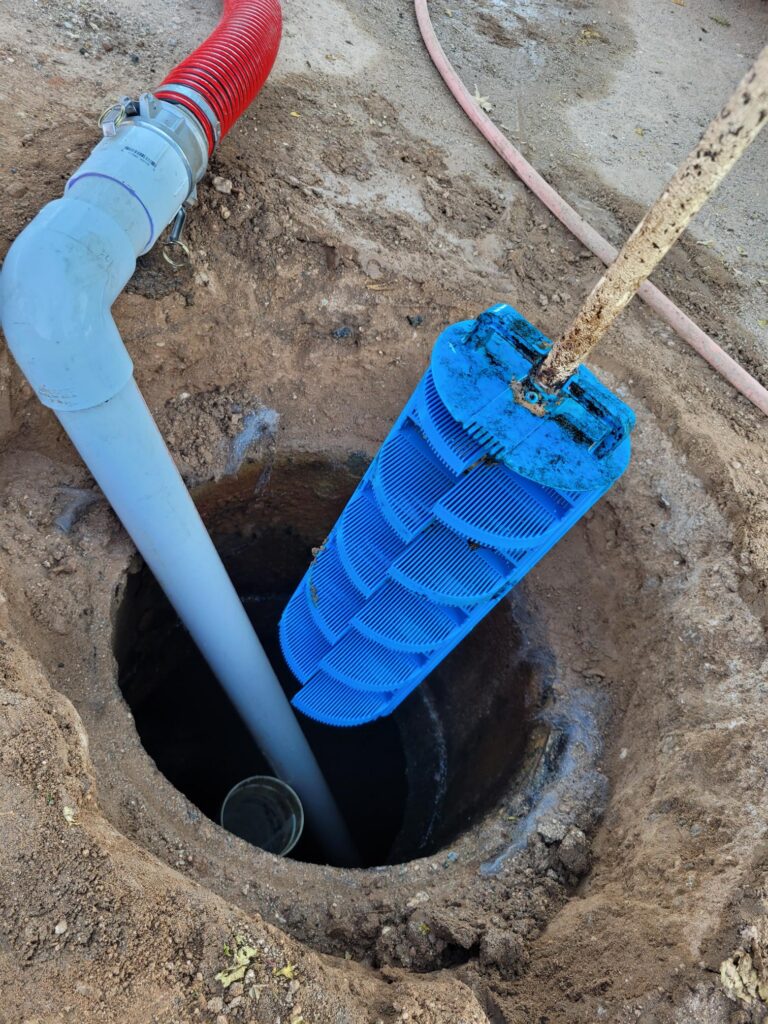 Blue submersible pump positioned in a dirt hole with a red flexible hose nearby on a construction site